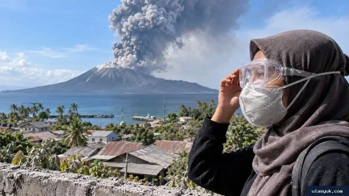 Warga mengenakan masker dan kacamata pelindung dengan latar gunung berapi yang mengeluarkan abu di kawasan pesisir.